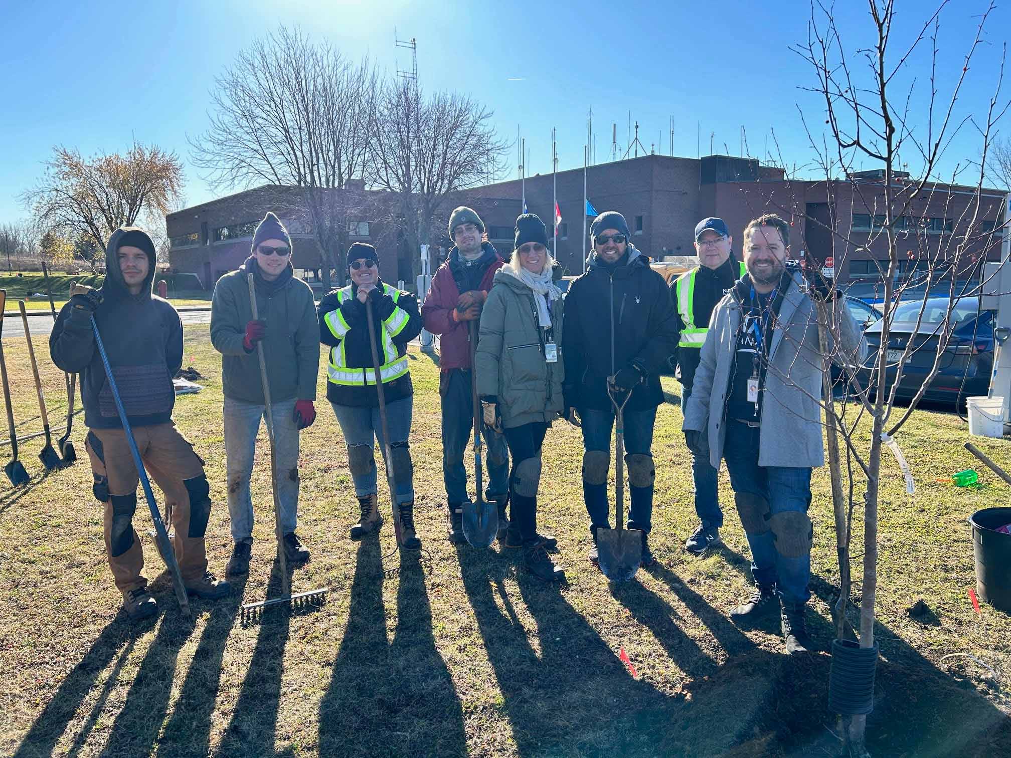 Group of people outdoors posing with shovels after planting a tree on a sunny day.